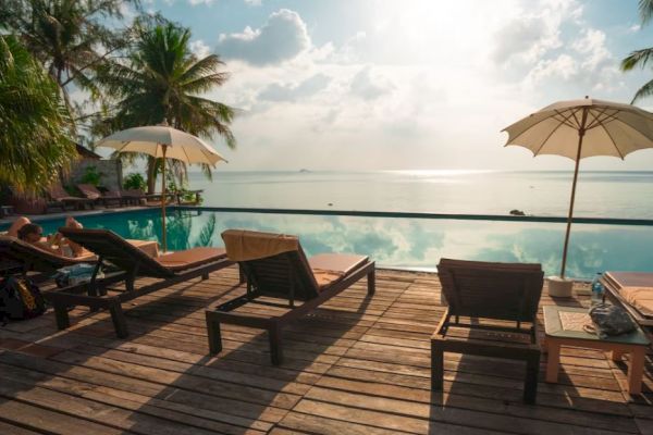 Sunlit poolside with lounge chairs, umbrellas, palm trees, and ocean view. Calm and relaxing atmosphere.