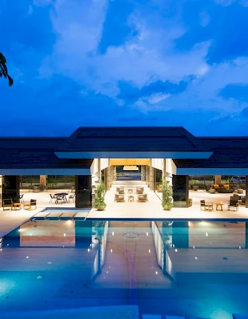 A modern, luxurious building with a large illuminated pool reflecting the architecture, set against a rich twilight sky.