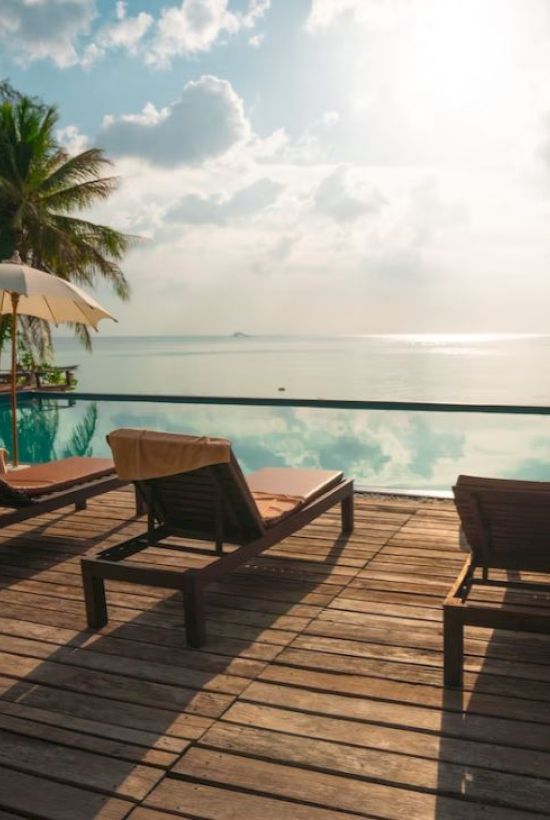 A serene poolside with lounge chairs and umbrellas overlooking a calm sea, surrounded by palm trees under a partly cloudy sky.