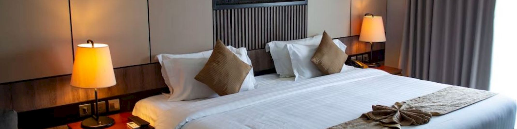 A cozy hotel room with a large bed, decorative pillows, and lamps on side tables, featuring warm lighting and a modern design.