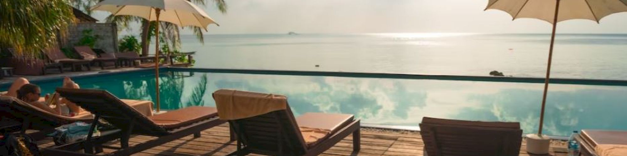 A serene poolside scene with lounge chairs, umbrellas, palm trees, and a view of the ocean under a partly cloudy sky is shown.