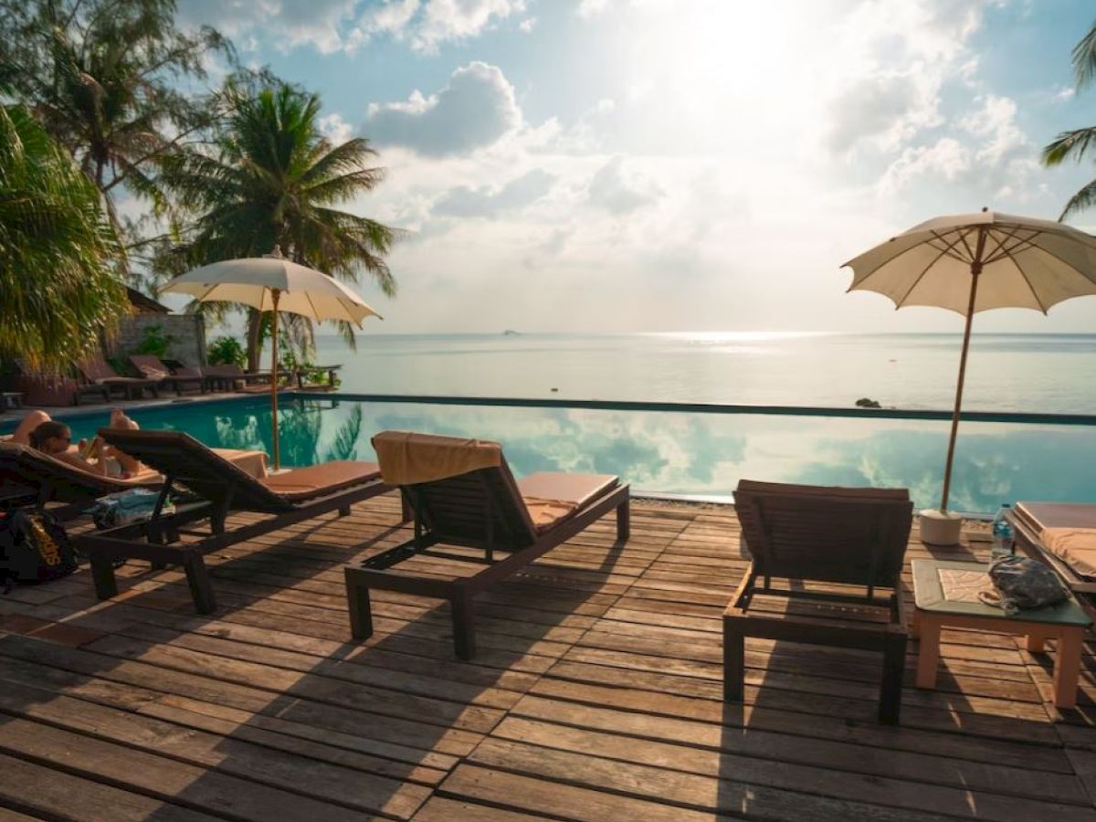 A serene poolside view with lounge chairs, umbrellas, palm trees, and a vast ocean under a bright sky.