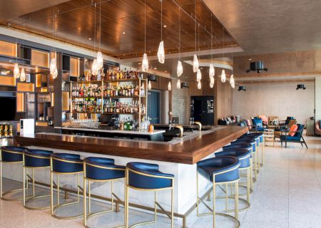 A modern bar with stylish seating, illuminated shelves stocked with bottles, pendant lights, and a cozy lounge area in the background.