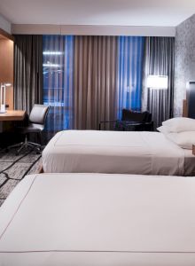 The image shows a modern hotel room with two beds, a TV, a work desk, and stylish lighting, featuring a patterned carpet and wall.