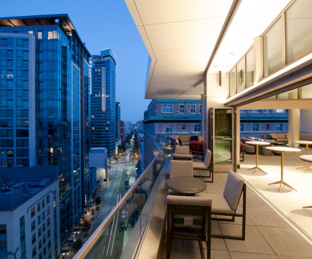 A modern rooftop patio with seating overlooks a cityscape at dusk, featuring tall buildings and illuminated streets.