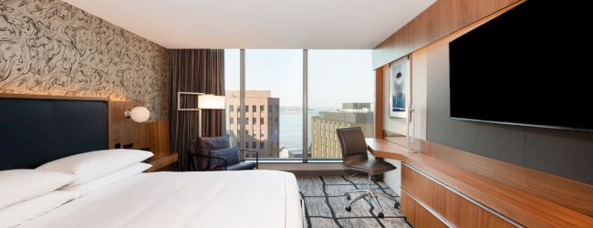 A modern hotel room with a large bed, wall-mounted TV, desk, and a view of buildings and water through large windows.