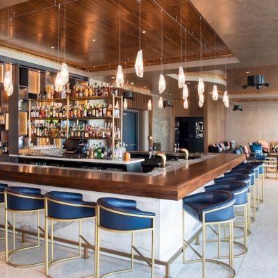 The image shows a modern bar with blue bar stools, pendant lights, and a variety of bottles on display, surrounded by a cozy seating area.