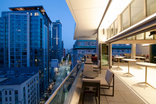A modern urban balcony view with outdoor seating, overlooking a city street and tall buildings at dusk.