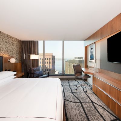 A modern hotel room features a large bed, a desk with a chair, a flat-screen TV, and a window with a city view in the background.
