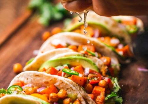 A hand squeezes lime over tacos filled with chickpeas, avocado, cilantro, and diced vegetables, arranged on a wooden board.