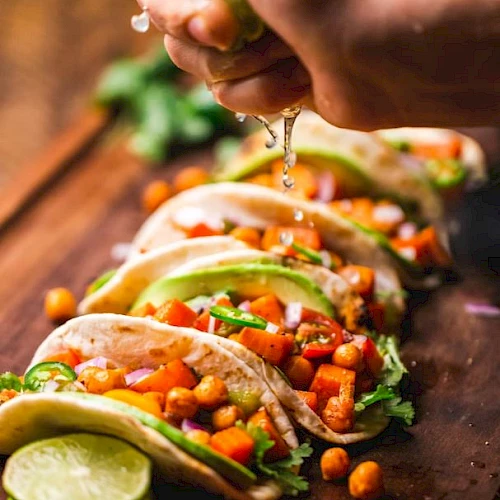 A hand squeezes lime over tacos filled with chickpeas, avocado, cilantro, and diced vegetables, arranged on a wooden board.