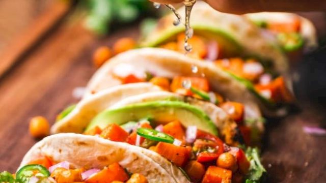 A hand squeezes lime over tacos filled with chickpeas, avocado, cilantro, and diced vegetables, arranged on a wooden board.