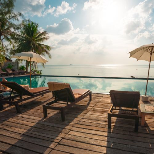 A serene beach scene with lounge chairs, umbrellas, and a pool overlooking the ocean at sunrise or sunset, surrounded by palm trees.