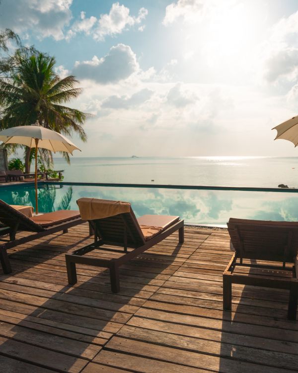 A serene beach scene with lounge chairs, umbrellas, and a pool overlooking the ocean at sunrise or sunset, surrounded by palm trees.