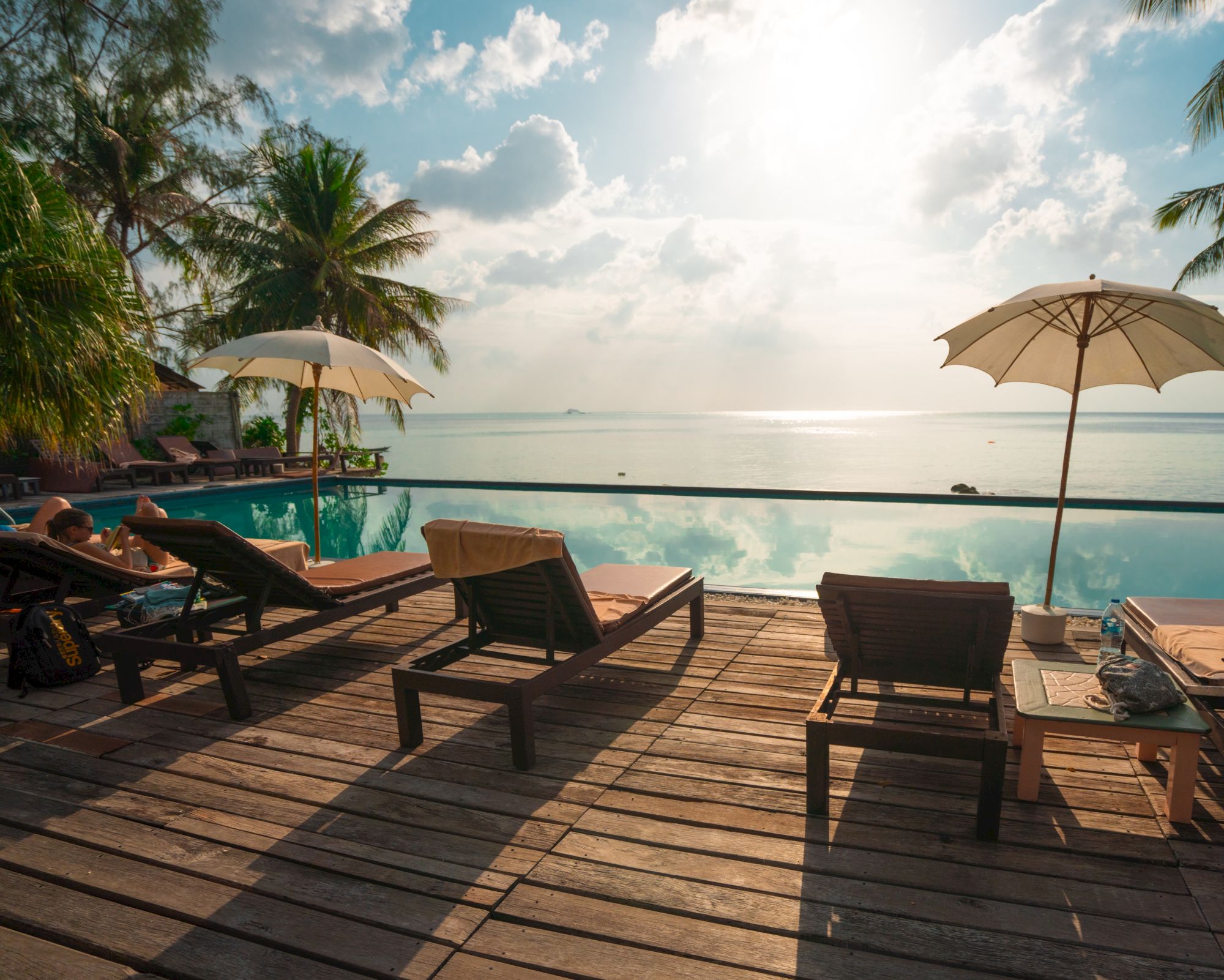 A serene beachfront scene with lounge chairs, umbrellas, palm trees, and a view of the sea under a bright sky reflected in an infinity pool.