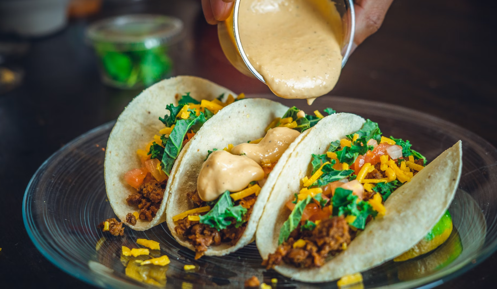 Three tacos with meat, cheese, tomatoes, and greens on a plate are being topped with a creamy sauce.