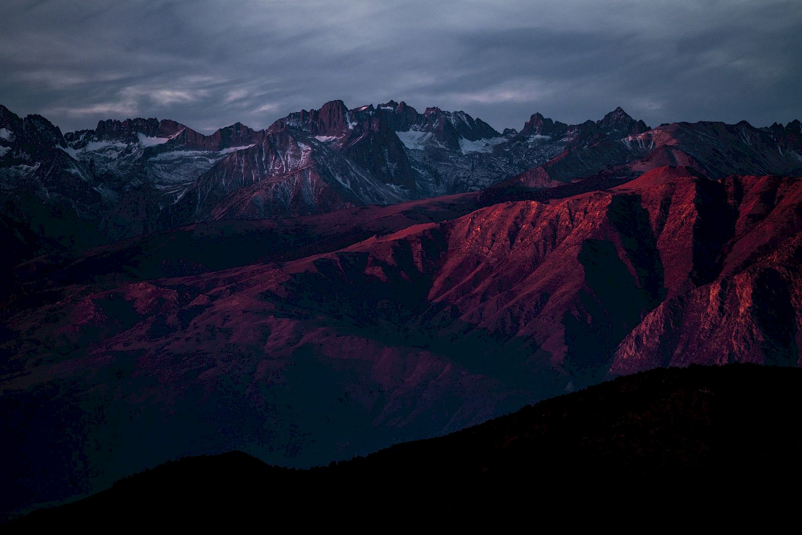 A mountain range under a dramatic sky, with peaks illuminated by red light during sunset, casting deep shadows across the landscape.