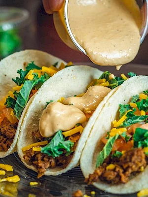 Three tacos filled with meat, cheese, lettuce, and tomatoes are being topped with a creamy sauce on a plate.