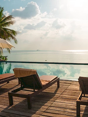 Poolside view with lounge chairs, umbrellas, and palm trees overlooking a calm sea under a cloudy sky at sunset or sunrise.