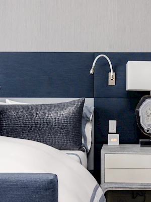 A modern bedroom features a bed with blue accents, a nightstand with a decorative lamp, and a wall-mounted reading light.