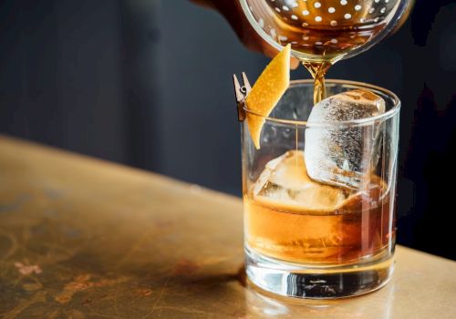 A cocktail being poured into a glass with ice and garnished with a twist of lemon peel clipped to the rim.