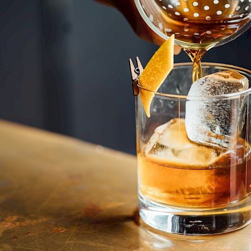 A cocktail being poured into a glass with ice and garnished with a twist of lemon peel clipped to the rim.