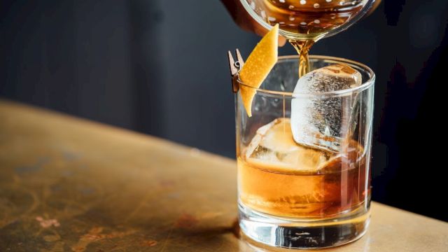 A cocktail being poured into a glass with ice and garnished with a twist of lemon peel clipped to the rim.