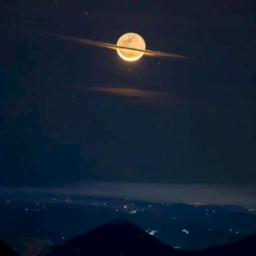 A full moon appears with cloud rings resembling Saturn's rings, over a mountainous landscape with distant city lights visible.
