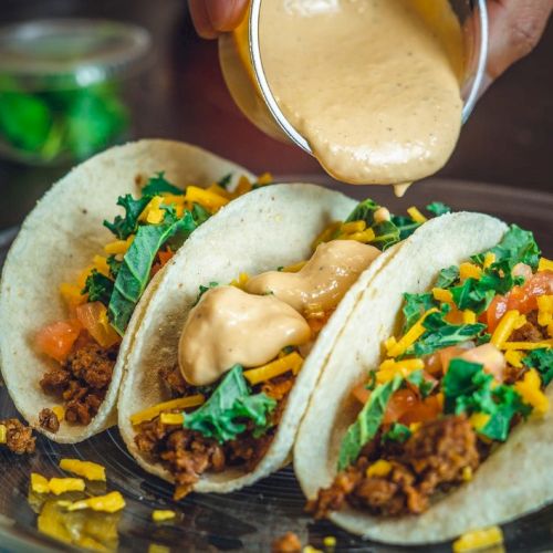 Three tacos filled with ground meat, cheese, greens, and tomatoes being topped with a creamy sauce on a plate.