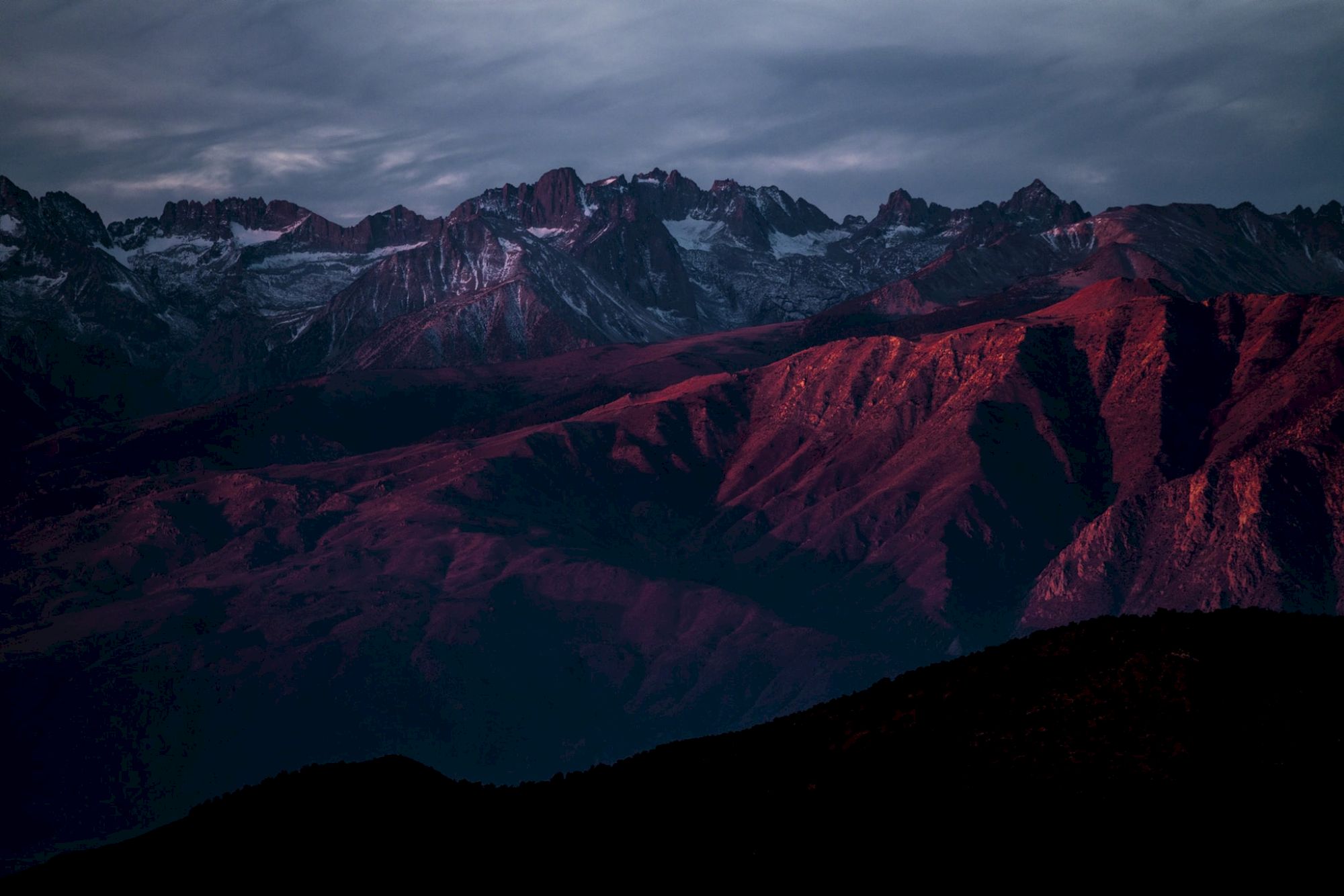 A mountain range bathed in a reddish sunset glow under a cloudy sky.