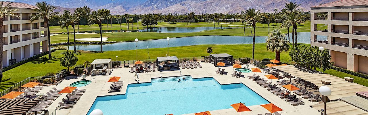 A resort pool area with loungers and orange umbrellas, surrounded by buildings, a golf course, and mountains in the background.