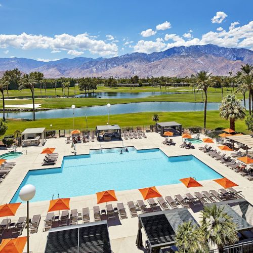 A resort pool area with loungers and orange umbrellas, surrounded by buildings, a golf course, and mountains in the background.