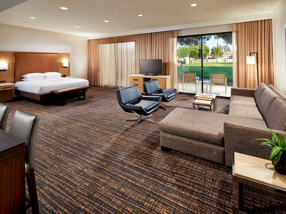 A spacious hotel room with a bed, sofa, chairs, TV, and large window overlooking a green landscape, offering a cozy and comfortable setting.