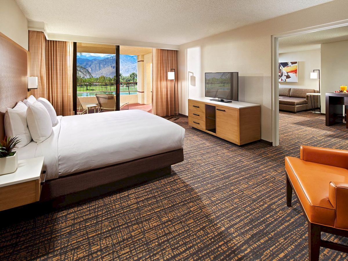 A spacious hotel room features a large bed, a sitting area with a TV, and a view of mountains through glass doors.