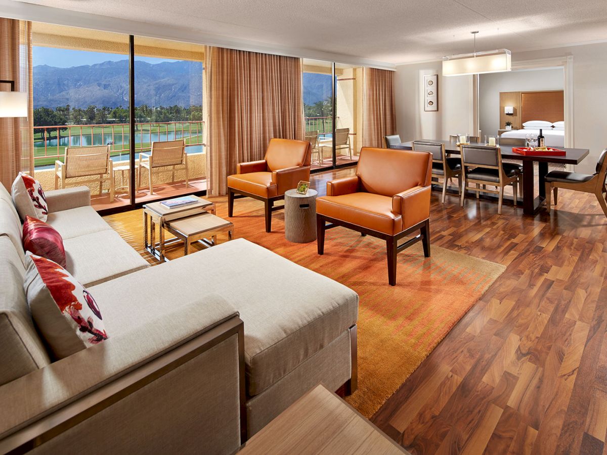 The image shows a modern living room with sofas, armchairs, dining area, wood floor, and large windows overlooking a scenic view.