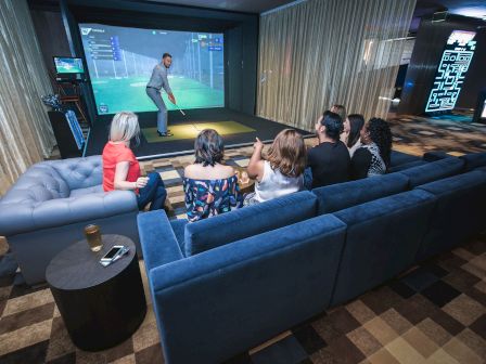 A group of people sits on sofas watching a man play in a golf simulator room, with a drink and a phone on a table nearby.