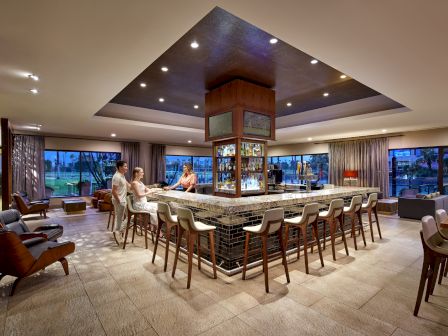 A hotel bar with a central island, surrounded by barstools, ambient lighting, and people conversing, with large windows in the background.