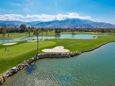 A scenic golf course with a lush green, water hazards, palm trees, and mountains in the background under a clear blue sky.