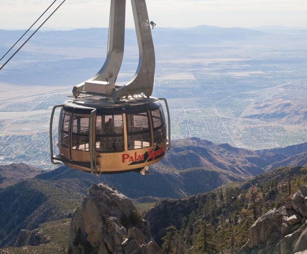 A rotating tram car is suspended over a mountainous landscape, with a view of the valley below and the text "Palm Springs" visible.