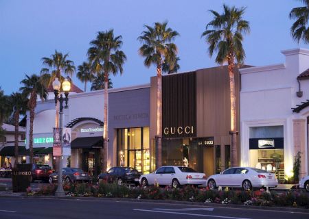 A stylish street with luxury stores like Gucci and palm trees, lined with parked cars under evening lighting, creating an upscale atmosphere.