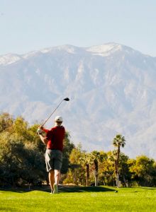 A person is playing golf on a green course with mountains in the background, under a clear blue sky.