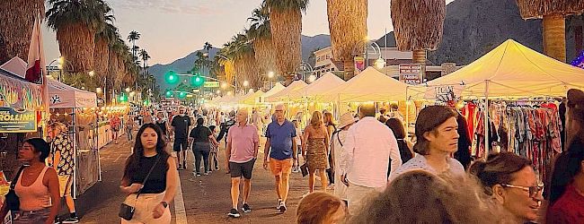A lively street market scene with people walking among vendor tents, palm trees lining the path, and mountains in the background.