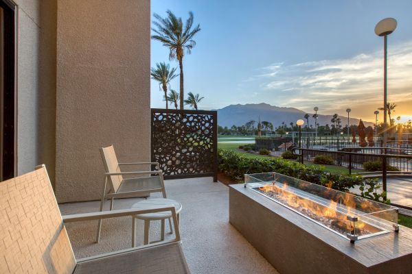 A patio with two chairs and a fire pit overlooks a scenic view with palm trees, a golf course, and mountains at sunset.