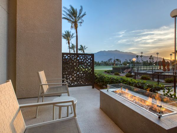A patio with two chairs and a fire pit overlooks a scenic view with palm trees, a golf course, and mountains at sunset.