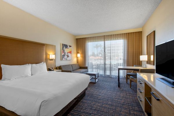 A hotel room with a bed, sofa, desk, TV, and window with curtains, featuring modern decor and neutral tones.