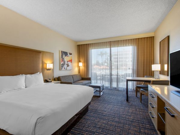 A hotel room with a bed, sofa, desk, TV, and window with curtains, featuring modern decor and neutral tones.