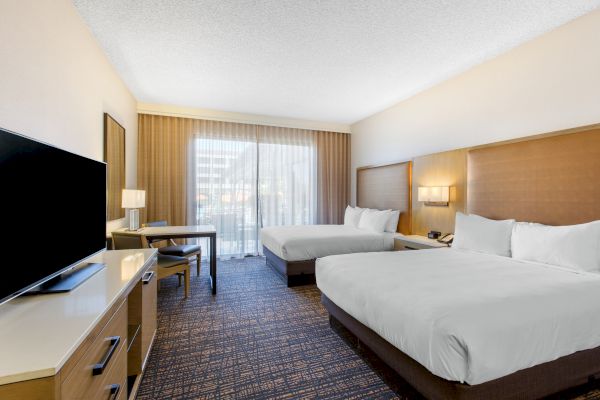A hotel room with two beds, a flat-screen TV, a desk, and a window with sheer curtains for natural light.