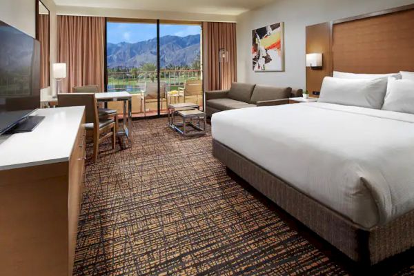A hotel room with a bed, sofa, desk, TV, and a view of mountains through a large window.