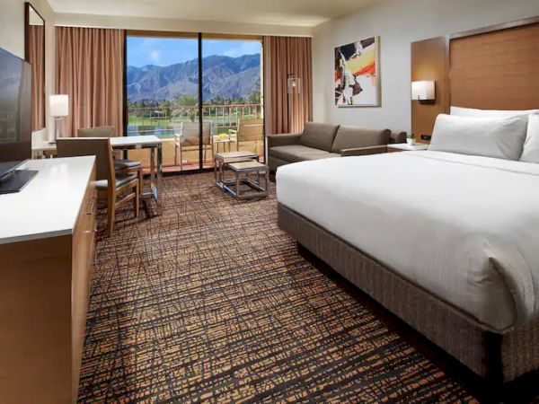 A hotel room with a bed, sofa, desk, TV, and a view of mountains through a large window.