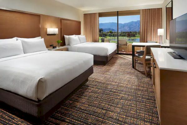 A hotel room with two beds, a TV, desk, and large window showing a scenic view of mountains and greenery.